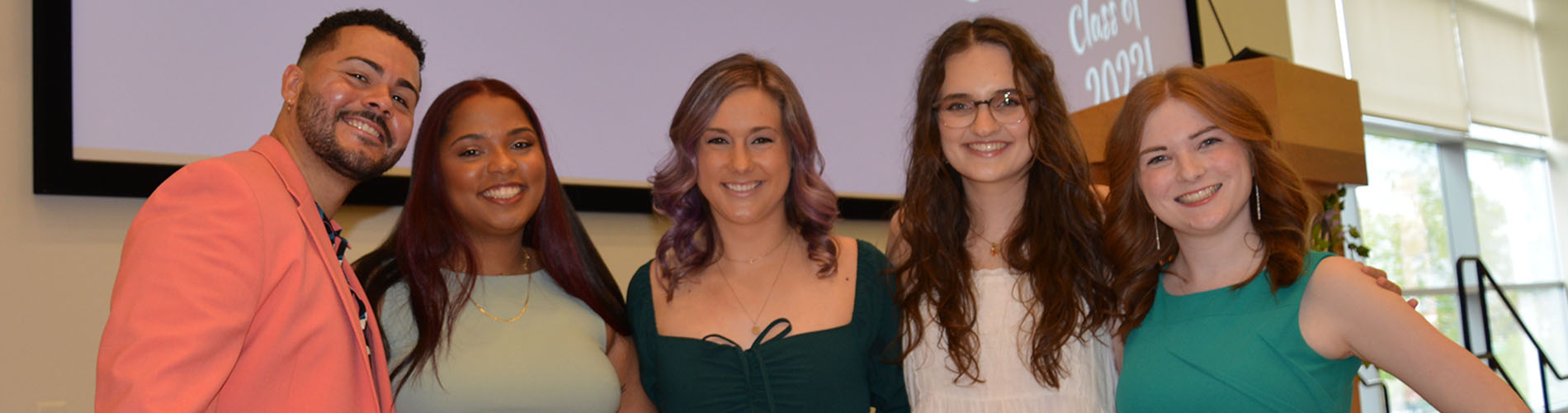 Five graduate students stand in a line and smile at the camera during a recognition ceremony.